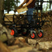 Person pushing a black superhandy utility wagon 48v with red wheels through a forest, carrying wooden planks.