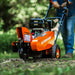 An orange and black SuperHandy stump grinder with a 9HP engine and a 12-inch cutting blade. Bottom Front view outdoors in use.