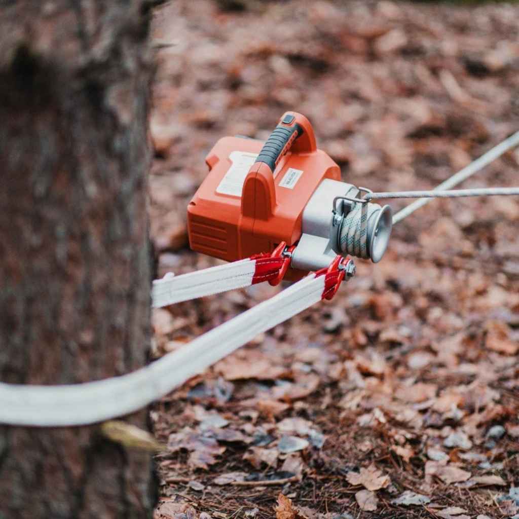 Red and silver superhandy capstan winch attached to a tree with a rope, on a forest floor.