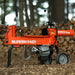 Orange SuperHandy log splitter 14 ton in a forest setting with stacked logs.